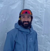 Alex wearing a blue ski coat, ski goggles, and smiling with the walls of a snowpit behind him.